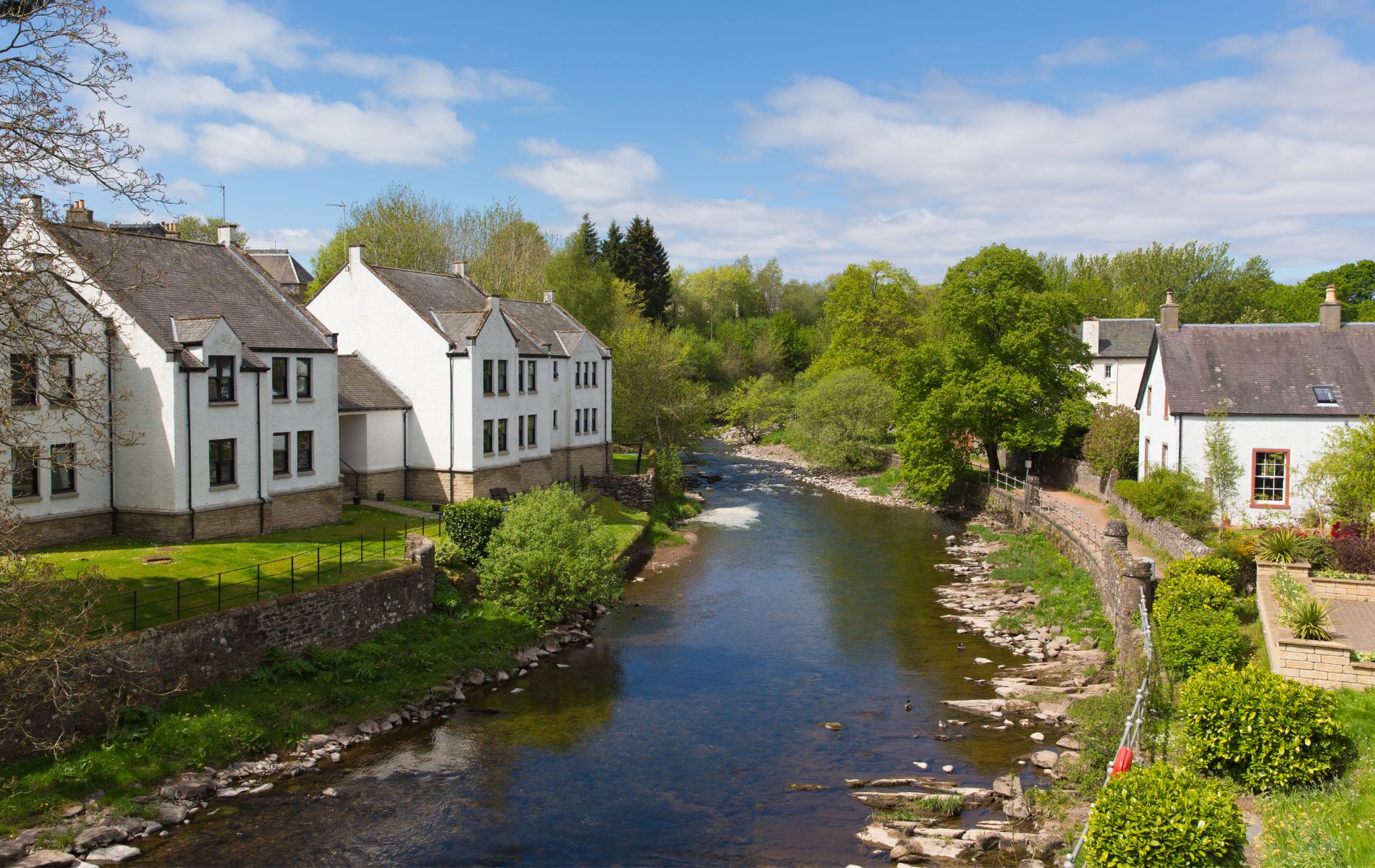 Riverside village with white houses along a calm river, green trees, and a bright blue sky on a sunny day.